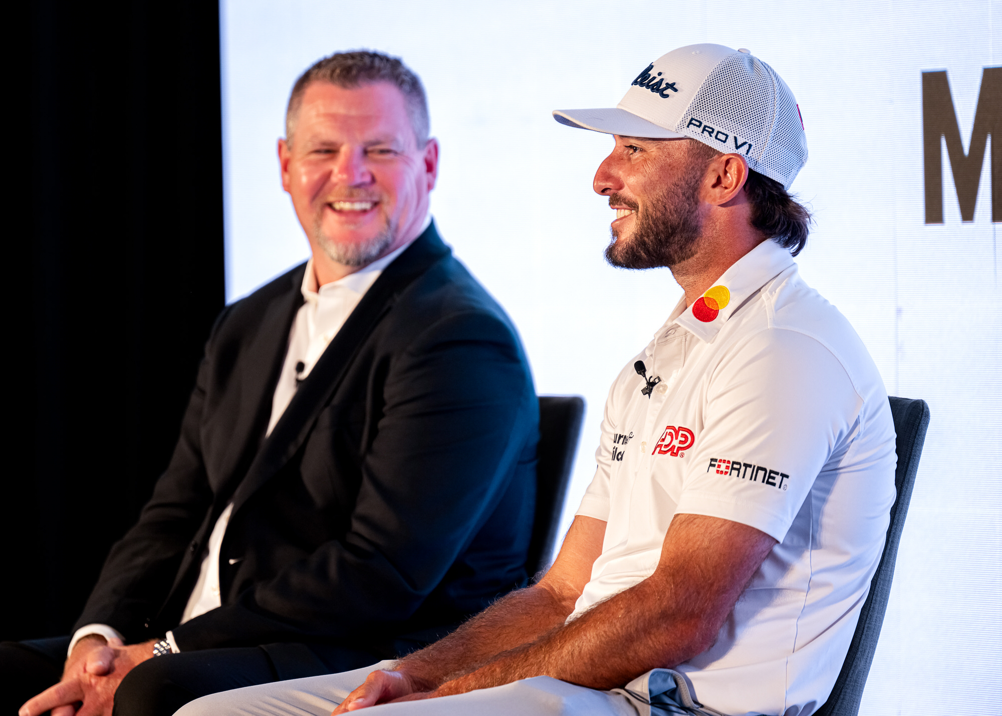 Fireside chat on stage between a host and a guest in a golf cap