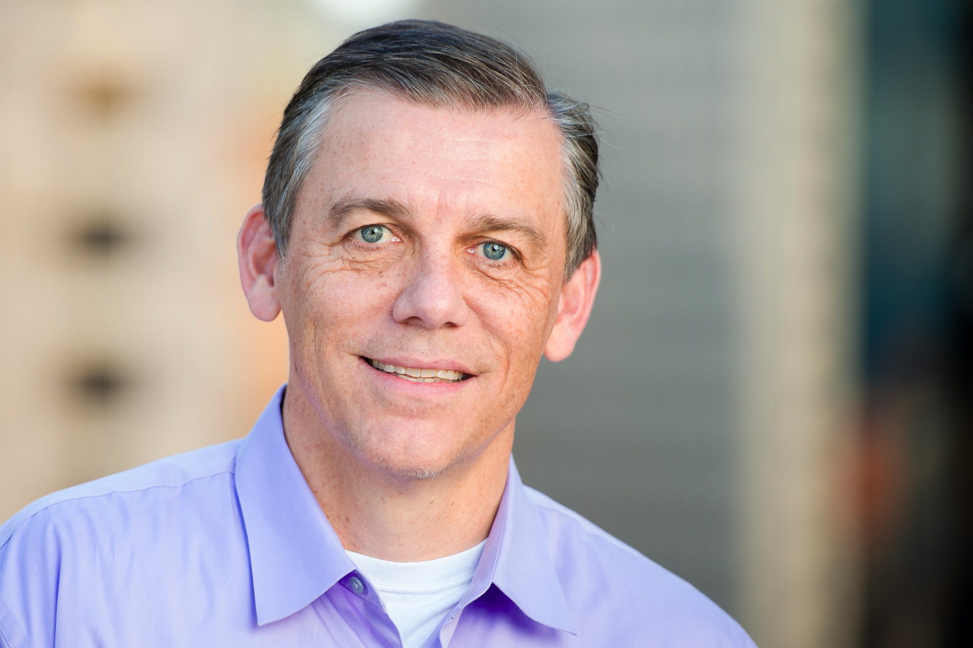 Corporate headshot of a smiling man in a lavender shirt, city buildings behind
