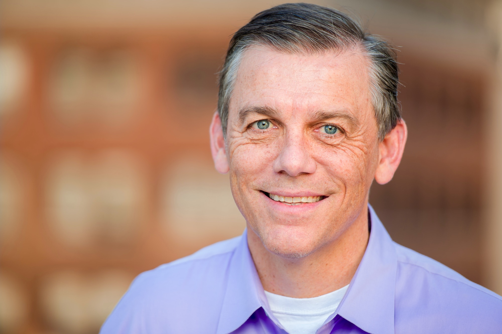 Corporate headshot of a man in a lavender shirt against a warm brick backdrop